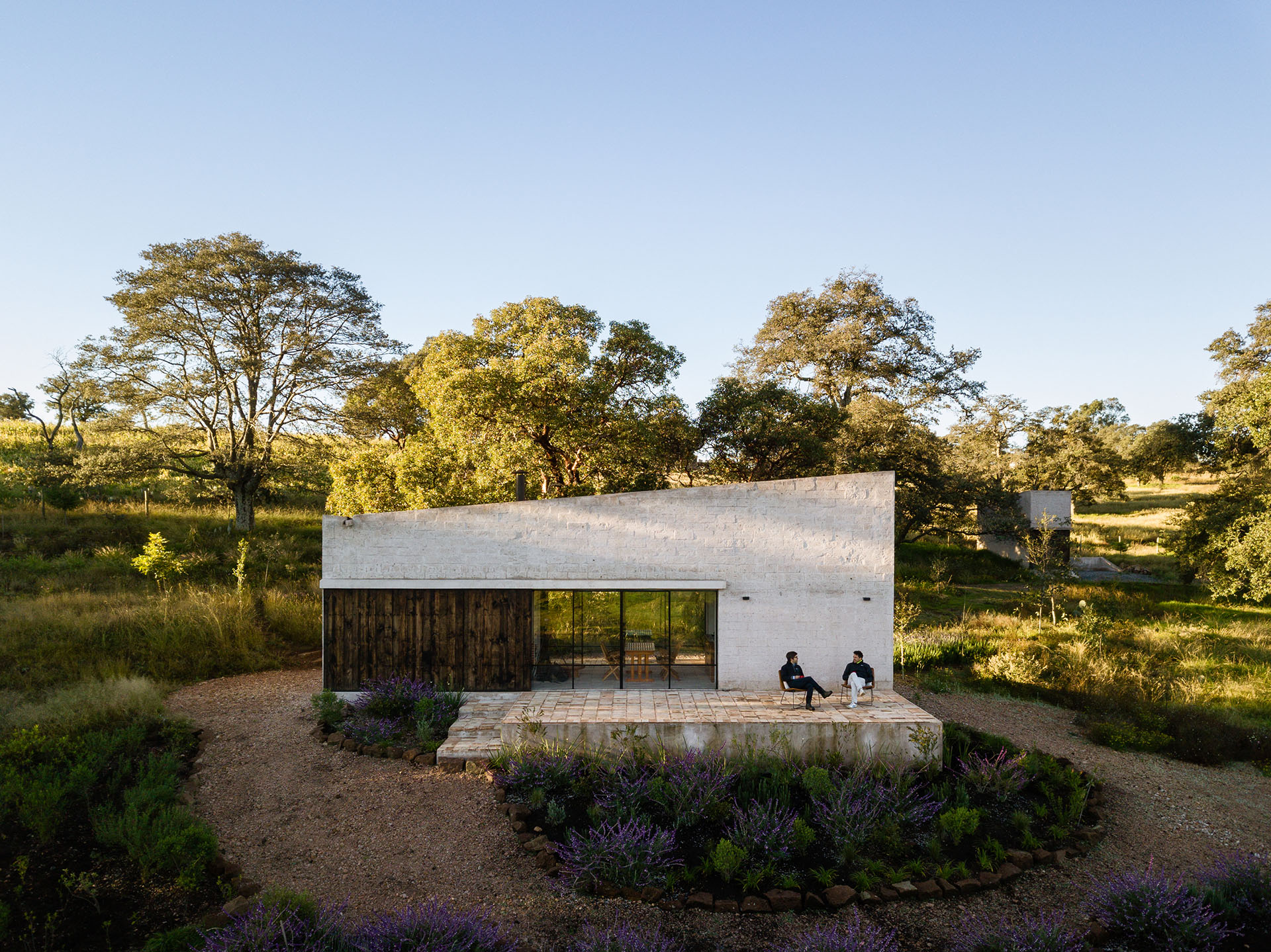 Casa rectangular de piedra clara con fachada acristalada en un paisaje arbolado.