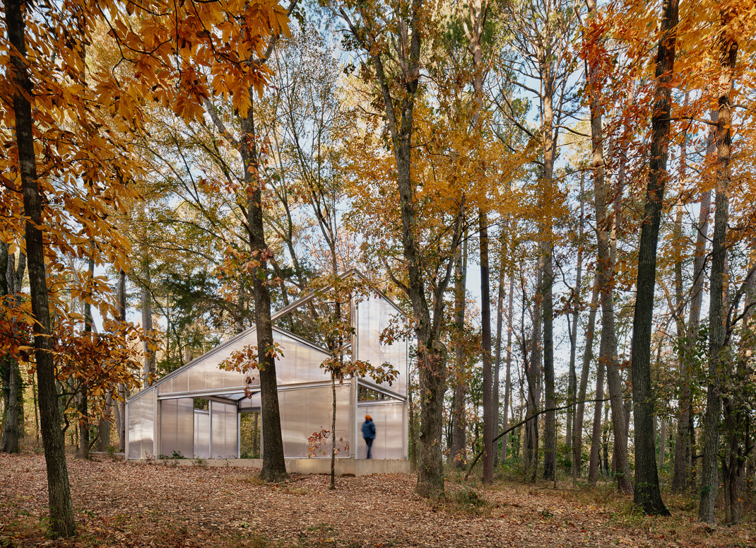 Cabaña triangular de Infinite Openness en un bosque otoñal, con una persona en la entrada.