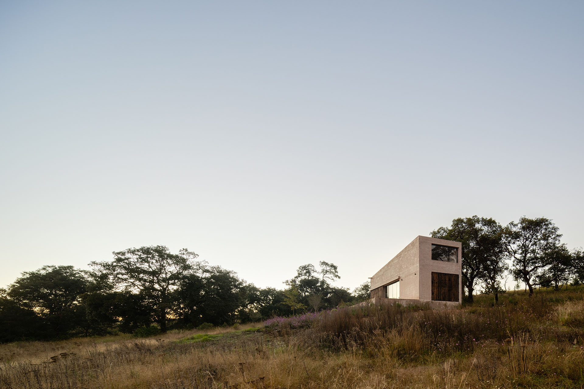 Casa compacta de piedra clara sobre una ladera de pasto seco al atardecer.