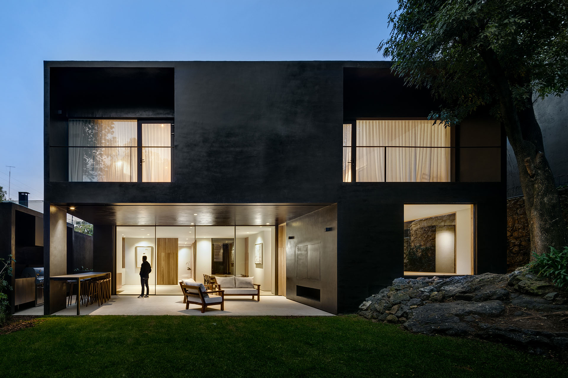 Casa negra iluminada al anochecer, abierta hacia un jardín con rocas.