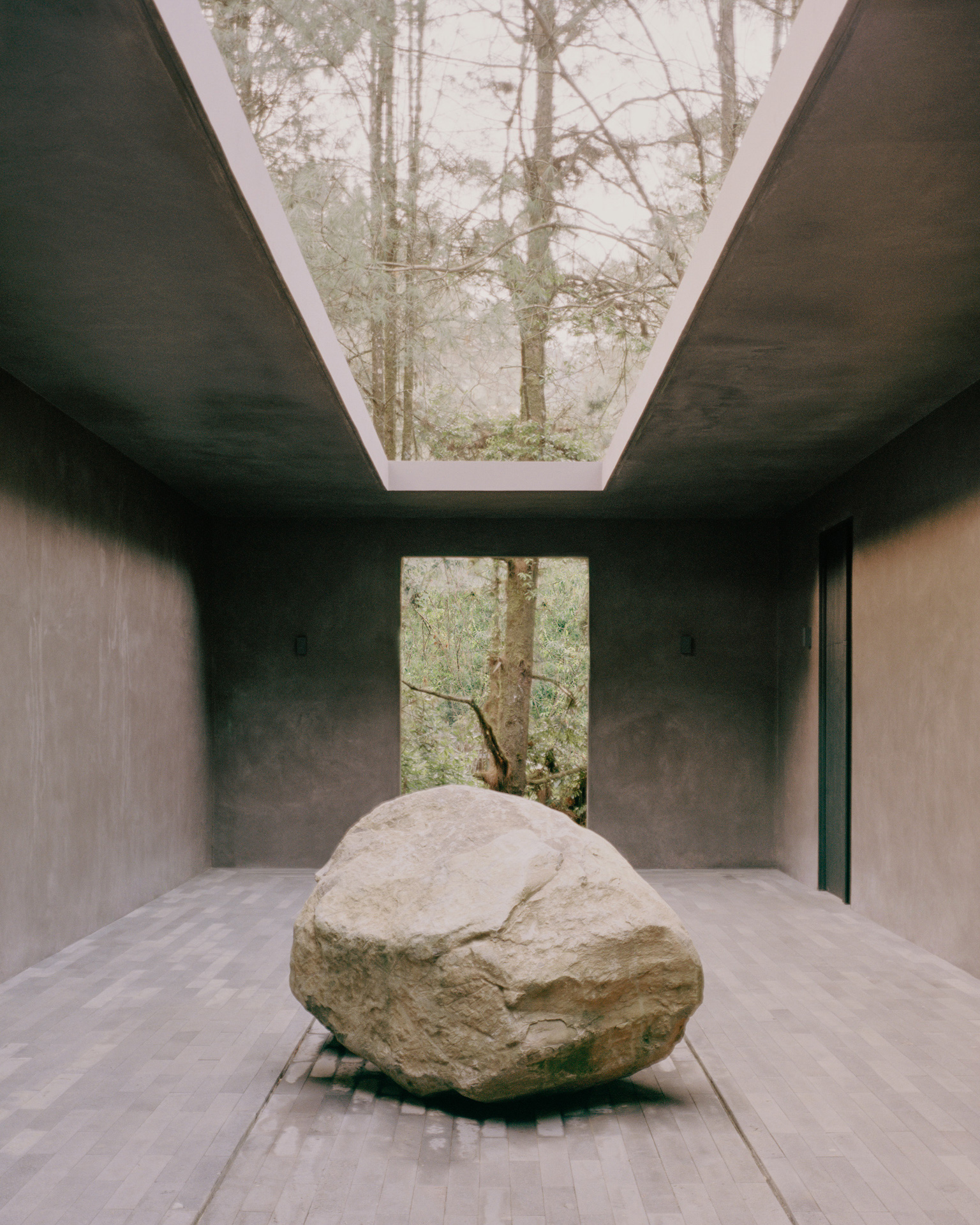 Gran roca colocada en una sala de concreto con abertura cenital y muro de piedra.